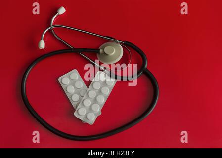 Close-up of a medical stethoscope and blister packs with pills shaped like the letter V on a bright red background, concept of victory over illness, t Stock Photo