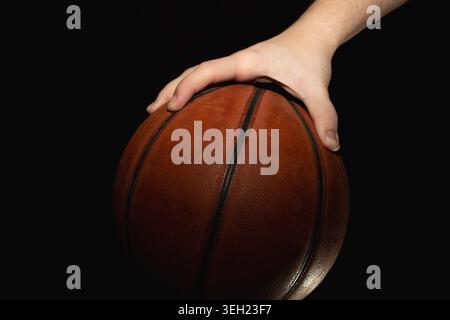 Hand resting on worn street basketball of dark brown color against black background. Leather surface is scuffed, visible abrasions and faded areas, bl Stock Photo
