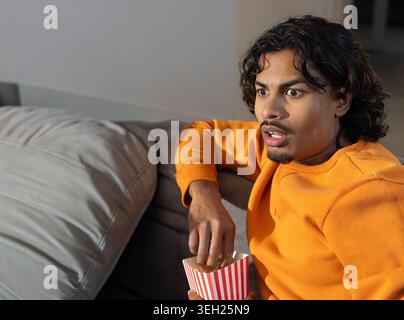 Indian man sitting on gray sofa at home wearing orange sweatshirt reaching into popcorn, copy space Stock Photo