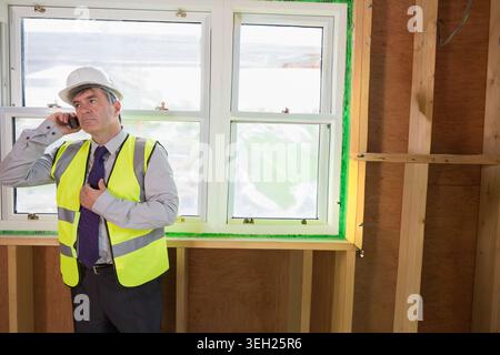 Mature man wearing hard hat, yellow vest, standing by sealed window holding phone and adjusting tie Stock Photo