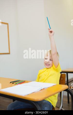Child sitting at desk in classroom, wearing yellow tee, raising arm with blue pencil, copy space Stock Photo