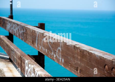 Vieste - Apulia - Italy - 08-29-2025: Close-up of a weathered wooden railing with faded graffiti, with clear turquoise seawater and a distant soft horizon in background. Stock Photo
