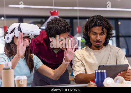 Diverse students collaborating at co-working table, interacting with white VR headset and tablet. Collaboration, teamwork, discussion, innovation, learning, workspace, modern Stock Photo