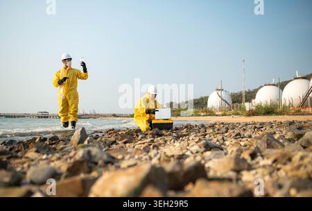 Scientists in yellow suits are collecting water samples from the coastline, flowing away from an oil refinery, before taking the water to the laboratory for further analysis Stock Photo