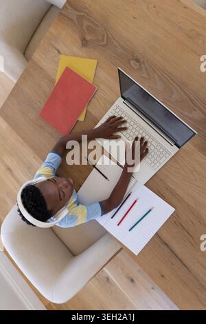 School-age African American boy sitting at wooden table wearing white headphones and using laptop. Learning, student, computer, blue, yellow, focus, minimalist Stock Photo