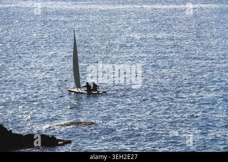 A small sailboat carrying two people glides across the calm blue sea. The minimalist marine scene features sparkling water, a distant perspective, and Stock Photo