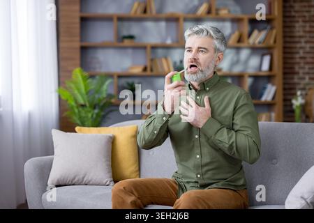Senior retired man experiencing shortness of breath from asthma or an allergic reaction, sitting on a sofa at home and using an inhaler to aid his breathing Stock Photo