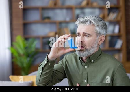Senior man with gray beard using an inhaler at home to relieve asthma or respiratory distress, showing shortness of breath, discomfort, and self-care treatment Stock Photo