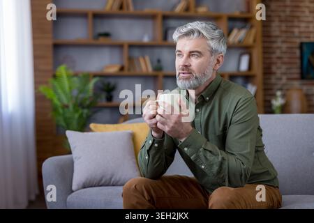 Mature gray-haired man relaxing on a sofa at home, holding a cup of coffee and gazing away in thoughtful reflection, enjoying a calm, restful break and quiet moment Stock Photo