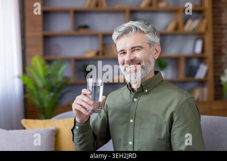 Mature man with a grey beard and a genuine smile promoting a healthy lifestyle while holding a glass of refreshing water, in his comfortable home environment Stock Photo