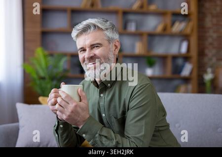 Happy mature man with grey hair and beard sitting comfortably on a couch, smiling at the camera while enjoying a warm cup of coffee in a relaxed home setting Stock Photo