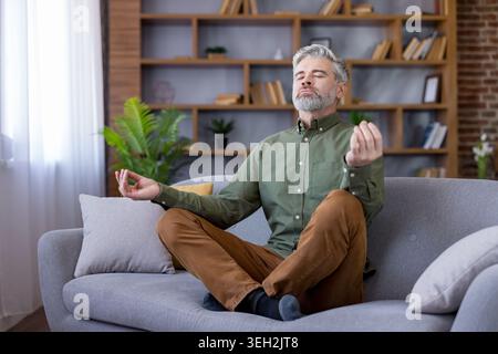 Mature man performs yoga meditation in lotus position on a comfortable couch at home, practicing mindfulness and relaxation while finding inner peace and mental balance Stock Photo
