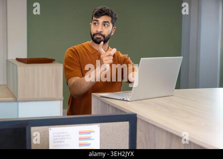 Indian man in rust-orange polo standing at reception counter pointing left and working on laptop Stock Photo