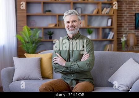 Mature man with gray hair and beard smiles confidently on a sofa with arms crossed, relaxed in a modern home office, conveying experience, expertise and remote work comfort Stock Photo