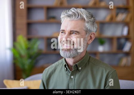 Middle-aged man with gray hair and beard smiles thoughtfully at home, relaxed and confident, envisioning future success and contentment while lounging in a casual living room setting Stock Photo