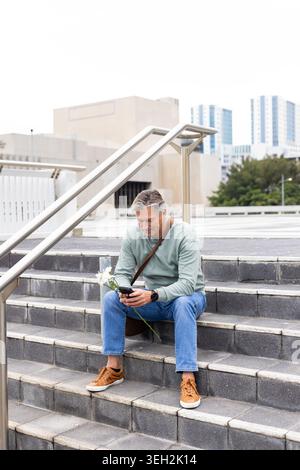 Mature man sitting on concrete steps at urban plaza holding white flowers and using smartphone Stock Photo
