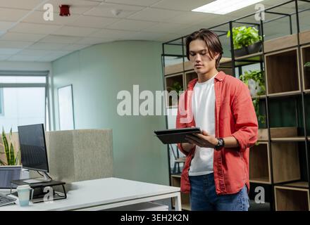 Asian man standing holding tablet, looking down in office wearing red shirt with laptop, copy space Stock Photo