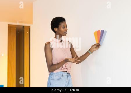 African American woman standing at home holding paint swatch fan to wall, in pink sleeveless top Stock Photo