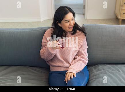 Woman in pink sweater sitting on sofa at home holding glass of red drink, looking right Stock Photo