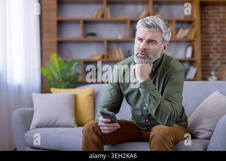 Mature man with gray beard sits on a sofa at home, holding a smartphone and resting his chin on his hand, pensive and contemplating future plans or recent thoughts Stock Photo