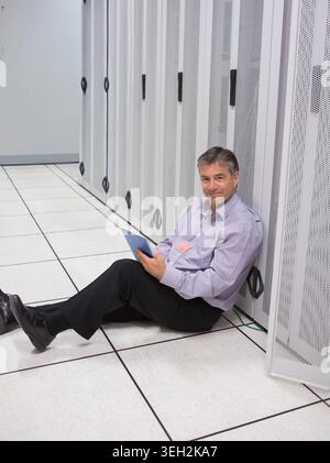 Middle-aged man sitting on floor in server room using tablet, wearing shirt, ID-badge, copy space Stock Photo
