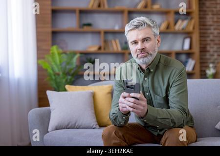 Mature man with stylish gray hair and beard sits on a modern sofa in a bright living room, holding a smartphone and engaging with digital life connected, relaxed, focused, contemporary Stock Photo