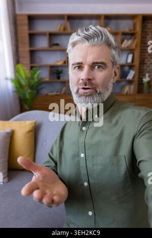 Middle-aged man with gray hair and beard actively talking during an online video call from his home office, communicating and explaining clearly from a first-person perspective Stock Photo
