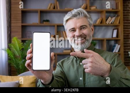 Happy senior man smiling directly at the camera, holding a smartphone with a blank white screen, and pointing towards it to emphasize a new application or communication Stock Photo