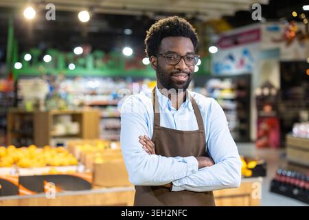Young man wearing an apron and glasses standing with arms crossed in a supermarket, representing entrepreneurship, customer service, and small business ownership Stock Photo