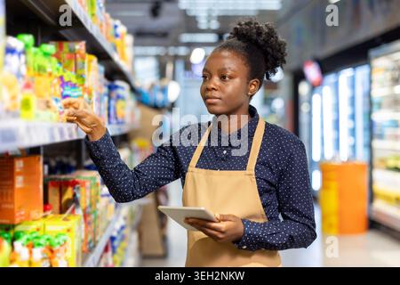 Young woman in apron and polo shirt stands in supermarket aisle checking shelf inventory with a digital tablet, scanning products and updating stock for efficient store management Stock Photo
