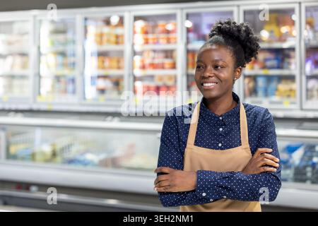 Young female shop owner standing confidently in her grocery store, wearing an apron and smiling, creating a welcoming and professional atmosphere for customers Stock Photo