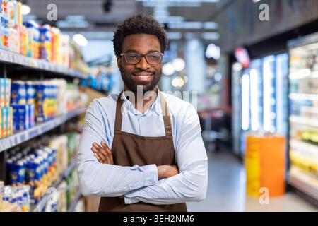 Young man wearing an apron and eyeglasses, smiling and looking at camera with arms crossed, working as a supermarket employee providing customer service in a grocery store setting Stock Photo