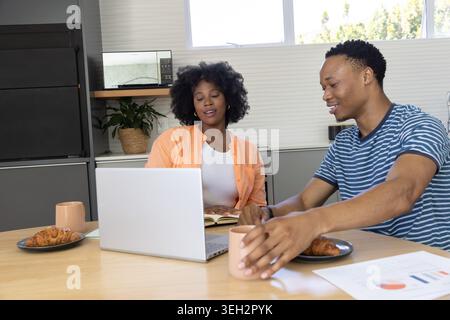 African American couple in orange shirt and blue striped tee working at kitchen table with laptop Stock Photo