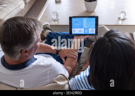Senior couple sitting on beige sofa at home using tablet showing smart-home controls, coffee table Stock Photo