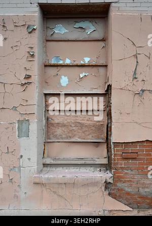 Vertical close up scene of a deteriorating tan painted wall with peeling paint, boarded window with rusty frame, exposed red brick Stock Photo