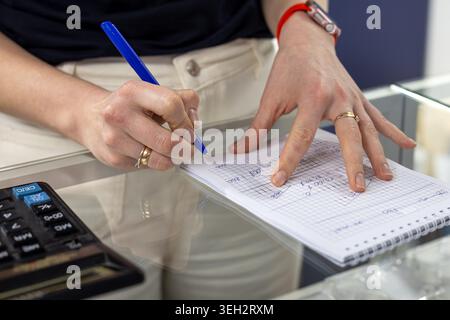 Hand Writing Notes With Calculator Nearby. Close-up of hands writing calculations in a notebook beside a calculator on a glass desk for budgeting or Stock Photo