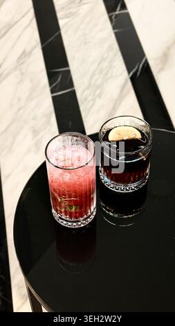 Two cocktail glasses on a black marble table, one filled with pink drink garnished with mint and the other with dark beverage and lemon slice Stock Photo