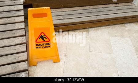 Yellow caution sign indicating wet floor placed on tiled surface near wooden steps, warning of potential slipping hazard in indoor environment Stock Photo