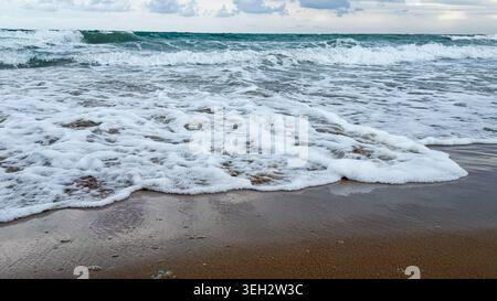 Waves crashing on sandy beach with foam and water reflections under cloudy sky, creating a serene coastal landscape in a natural setting Stock Photo