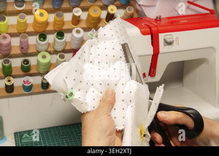 Person crafting textile item, using scissors to cut fabric. A sewing machine and colorful threads are visible in the background Stock Photo