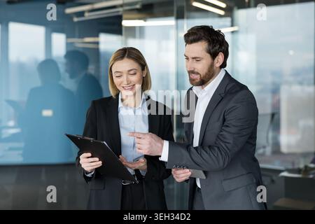 Two business colleagues, man and woman in professional attire, standing in a modern office, collaborating over a clipboard, discussing plans and reviewing documents with confident expressions Stock Photo