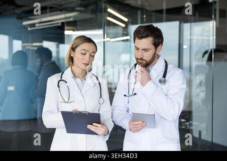 Two doctors wearing lab coats and stethoscopes are discussing patient data, one reviewing a clipboard and the other analyzing information on a digital tablet in a modern clinic setting Stock Photo