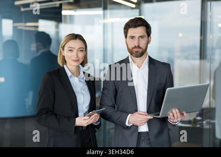 Two confident business partners standing in a modern office, a man holding a laptop and a woman holding a digital tablet, representing teamwork and corporate success Stock Photo