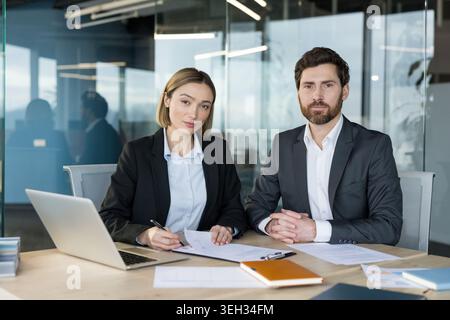Two confident business people discussing a project, reviewing documents, and working on a laptop at a contemporary desk, representing teamwork and professional collaboration Stock Photo