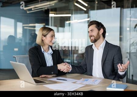 Two business professionals smiling and shaking hands over a desk with a laptop and documents, sealing a successful partnership or agreement in a modern office environment Stock Photo