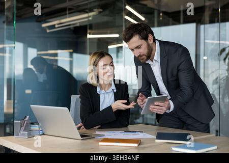 Two professional business colleagues collaborating on a project, analyzing data and discussing strategy using a digital tablet and laptop in a modern corporate office setting Stock Photo