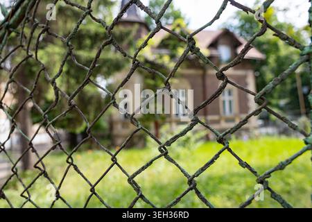 Old rusty metal wire mesh fence close-up with a blurred historic house and green yard in the background Stock Photo