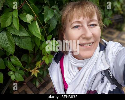 A woman smiles joyfully in a vibrant park, standing amidst rich green leaves on a sunny day in early autumn. Stock Photo