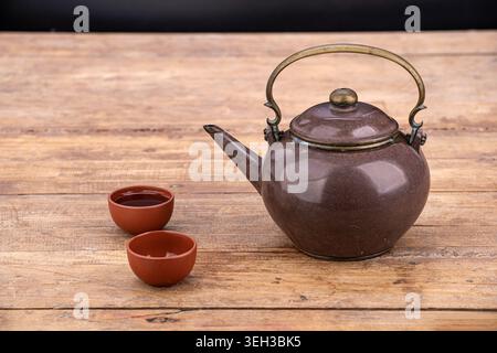 A traditional vintage brown glazed ceramic teapot with an ornate brass metal handle and lid knob is displayed alongside two small terracotta clay tea Stock Photo