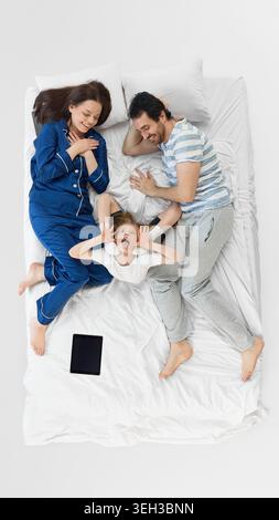 Playful daughter making funny face between parents on white bedding. Stock Photo